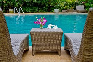 A photo taken from behind two wicker lounge chairs and table that has flowers on it overlooking a swimming pool in the background.