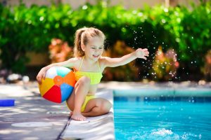 A young girl in a yellow bathing suit happily kneeling by the edge of a pool holding a colorful beach ball.