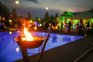 A close up of a fire feature with a pool and party goers in the background at night.