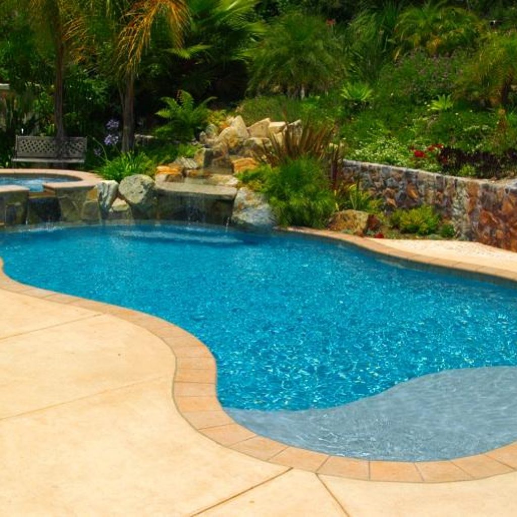 A freeform zero entry pool with a raised spa against a tropical landscape in a residential backyard.